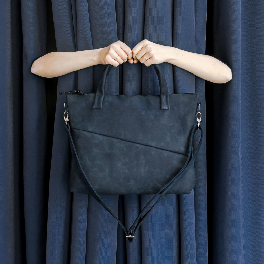 Two hands holding shoulder bag ANA with handles and shoulder strap in dark blue natural leather