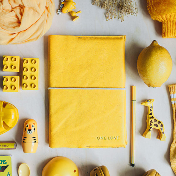 Mood Shot U-book cover EVE made of sustainable natural leather in curry with individual embossing in gold and cream colored closure band next to yellow toy