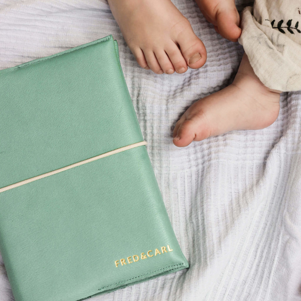 Baby feet next to U-booklet EVE as a sibling cover made of natural leather in mint with name embossing in gold and cream-colored closure band